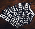 Five OCEAN CREST die-cut stickers fanned on dark wood.